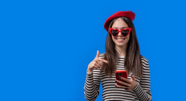 Smiling teenager girl pointing to smartphone, she advise mobile app, mobile operator. Young woman on blue background. Banner, copy space. High quality photo