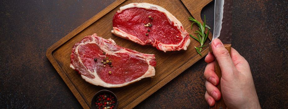 Male's hand holding knife over two raw uncooked meat beef rib eye marbled steaks on wooden cutting board and seasonings on dark rustic background from above, preparing dinner with meat