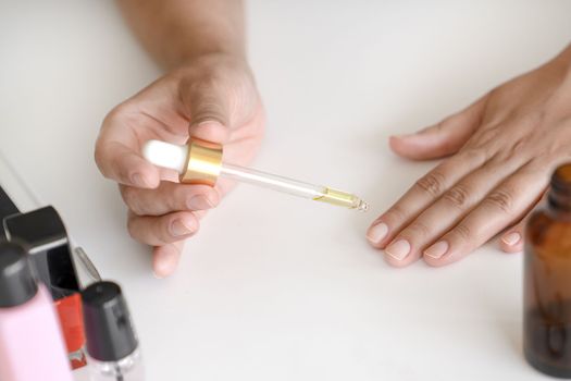 Woman holds pipette with oil to apply to her nails for treatment and strengthening of nails. Manicure accessories are next on table. Hands close-up, no face