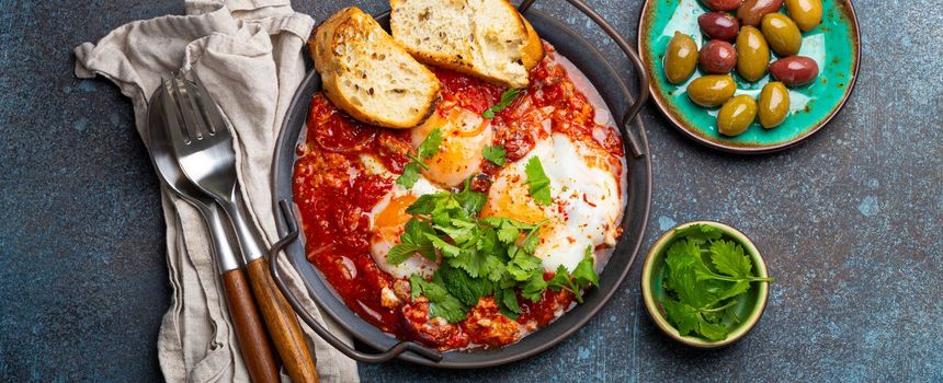 Middle Eastern and Maghrebi healthy dish Shakshouka made of eggs and tomato sauce served in pan with toasts, fresh cilantro and olives on rustic concrete background table from above