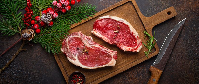 Two raw uncooked meat beef steaks on wooden cutting board with knife and seasonings on dark rustic background with Christmas festive decoration from above