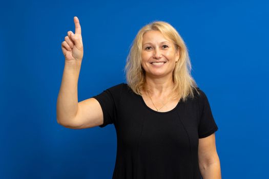 Smart proactive 40 years old woman in black t-shirt holding up index finger with great new idea isolated on blue colored background studio portrait. People lifestyle concept