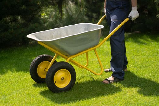 A young man with hands in gloves is carrying a metal garden cart through his beautiful green blooming garden. A professional gardener is carrying a wheelbarrow