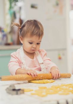 Adorable baby in an apron is rolling out the dough in the kitchen.