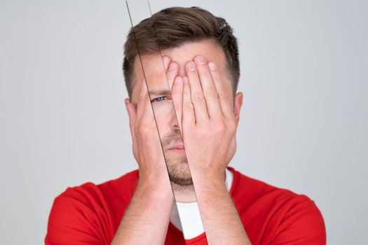 Young man covering his face by hand and looking at camera while standing against grey background. High quality photo