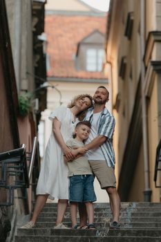 A full-length photo of a family which is hugging on the stairs in an old European town. A happy father, mother, and son are holding hands and having fun in the evening.