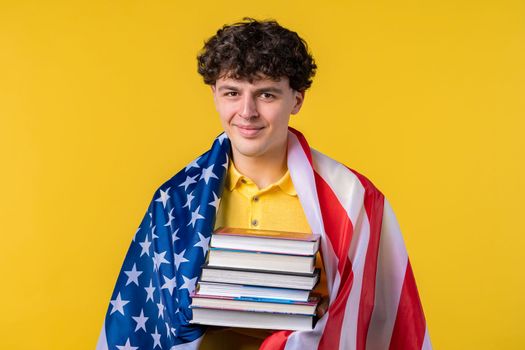American man student holds stack of university books from college library on yellow background. Happy guy smiles, he is happy to graduate in USA, education abroad concept. High quality photo