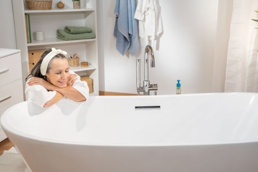 Beautiful middle aged woman in bathrobe leaning with arms crossed relaxing at luxury bathtub. Body care spa procedures antiage recreation skin care and beauty cosmetic concept.