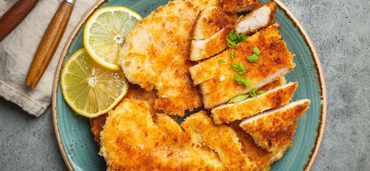 Crispy panko breaded fried chicken fillet with green salad and lemon cut on plate on gray rustic concrete background table with ketchup from above. Japanese style deep fried coated chicken breasts.