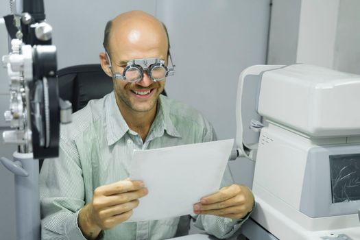 Smart handsome man sitting in optometrist cabinet having his eyesight checking, examining, testing with trial frame glasses by professional optician for new pairs of eyeglasses.