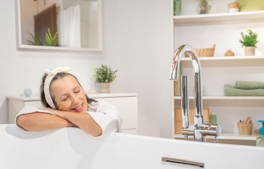 Smiling middle aged woman in bathrobe and headband leaning with crossed arms, relaxing with closed eyes at bathtub. Bodycare spa procedures antiage recreation skin care and beauty cosmetic concept.