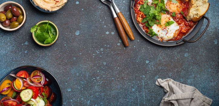 Middle Eastern traditional breakfast or brunch with eggs Shakshouka in pan with toasts, fresh vegetables salad, hummus and olives on rustic concrete background table from above, space for text