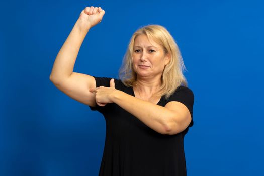 Confident caucasian blonde woman pointing finger at her arm muscle, demonstrating her strength and independence, women's rights, feminism. Indoor studio shot isolated on blue background.