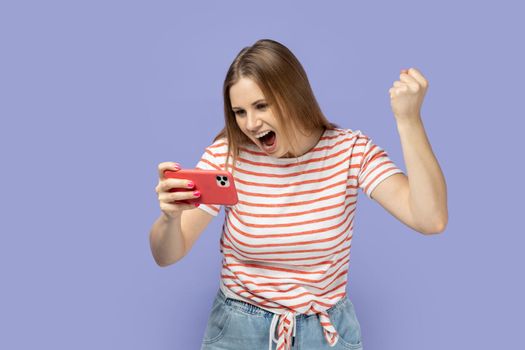 Portrait of excited smiling woman gamer wearing striped T-shirt playing game on smart phone, winning level, clenched fist, celebrating her victory. Indoor studio shot isolated on purple background.