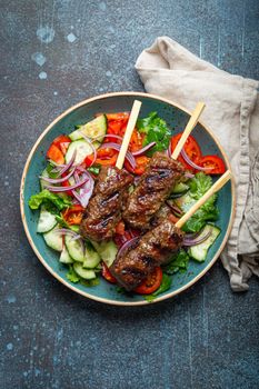 Grilled skewer meat beef kebabs on sticks served with fresh vegetables salad on plate on rustic concrete background from above. Traditional Middle Eastern and Turkish dish Kebab