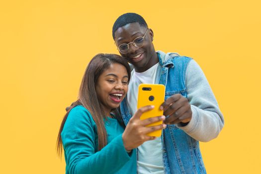 Excited African American couple looking at smart phone screen together isolated on yellow background. Joyful black man and woman using cellphone together, entertainment concept. High quality photo