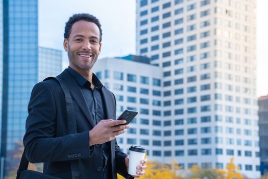 Portrait of successful black businessman in suit smiling looking at camera with financial buildings in background, smart phone and coffee cup. High quality photo