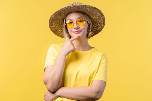 Portrait of young pretty blonde woman on summer yellow studio background. Confident sunny outfit with sunglasses and straw hat. High quality photo