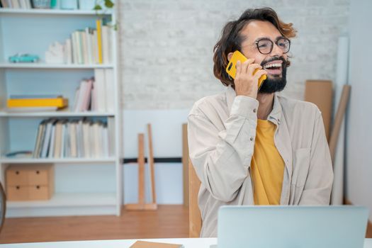 Young creative entrepreneur talking on the phone laughing smiling in his modern workspace at home - Freelance guy happy talking to client with white and yellow colors office with copy space. High quality photo