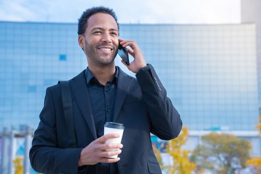 Successful African American businessman in suit smiling happy talking with smart phone with financial buildings in background and coffee cup. High quality photo
