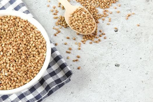 Buckwheat in a white bowl with wooden spoon, raw green buckwheat grain on napkin and light grey stone background, close up, top view.