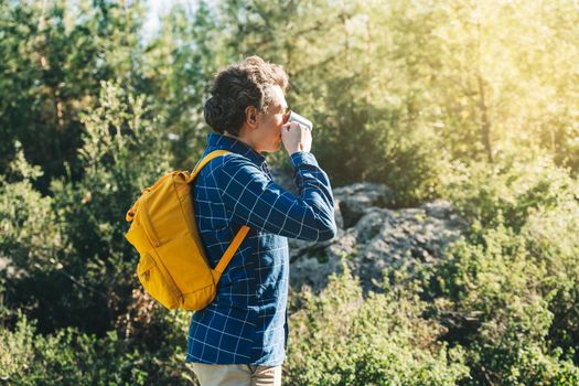 Adult male tourist with yellow backpack walking in forest. Young guy in casual clothes looking at view, drinking hot coffee or tea in metal cup while hiking in summer greenwood forest.