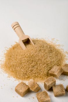 Brown cane sugar is also sprinkled in lumps on a light surface. Wooden scoop for bulk products. Selective focus. Vertical orientation, space for text.