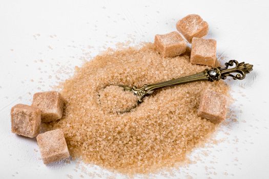 A pile of granulated cane sugar and pieces on a light surface with a small elegant teaspoon. Selective focus.