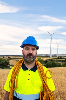 Vertical portrait of male maintenance worker standing in wind turbine farm field with climbing equipment: helmet and rope. Looking at camera. Engineering and renewable energies.