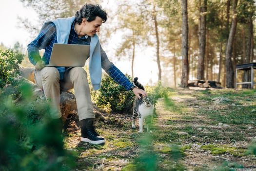Happy cheerful man with a laptop sitting outdoors on rock in nature forest park and pet cat. Freelancer with computer typing, blogging, browsing in vacation. Freelance, distance work concept.