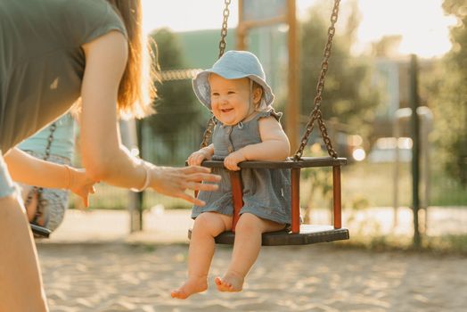 Toddler baby girl on a swing on the warm summer evening. Mother is swinging her young daughter on a sunny playground.