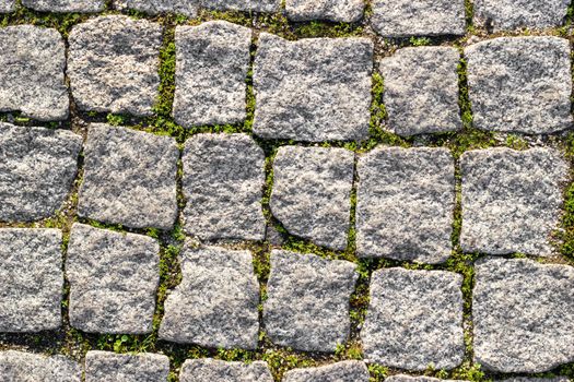 Texture of paved stone road with asymmetrical stones with sprouted grass at seams. Top view