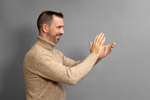Bearded hispanic man in profile wearing brown turtleneck feeling happy and successful, smiling and clapping hands, saying congratulations with a clap against copy space gray wall