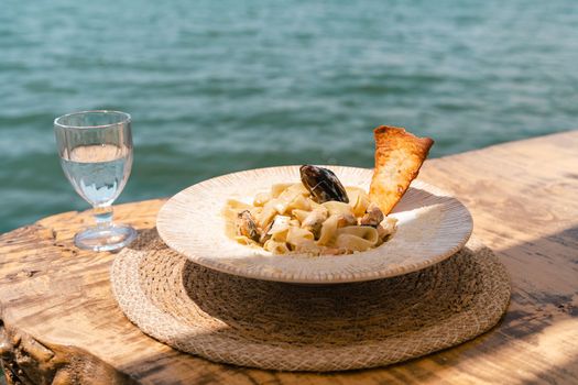 Seafood pasta spaghetti and a glass of water standing on a wooden table in outside cafe restaurant with scenery sea in the background. Copy blank space. Fish diving into noodles concept.