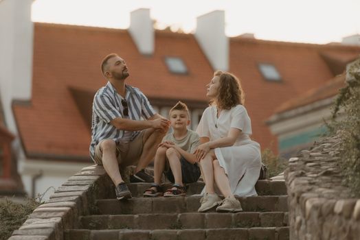 A family is relaxing on the stairs between roofs in an old European town. A happy father, mother, and son are having fun in the evening.