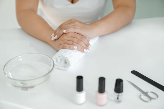 Women's hands with white manicure are lying on towel roller, waiting for varnish to dry, manicure accessories are lying on table next to it. Hands close-up, front view, without face