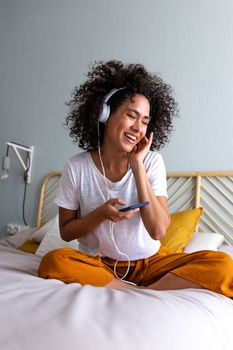 Happy young African American woman laughing listening to podcast. Relaxing listening to music with headphones and mobile phone in bedroom. Vertical image. Lifestyle concept.