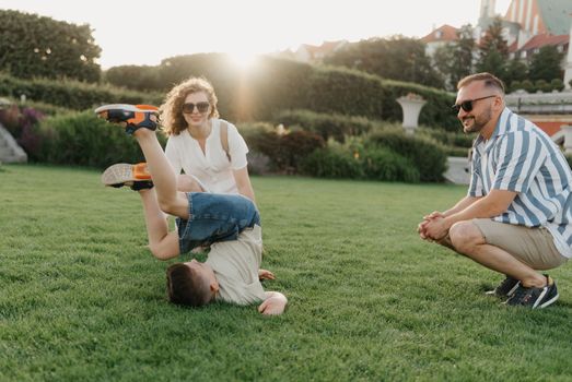 Father, mother, and son are having fun in the garden of an old European town. Happy family in the evening. A boy is showing somersaults to his parents in the background of the palace at sunset.