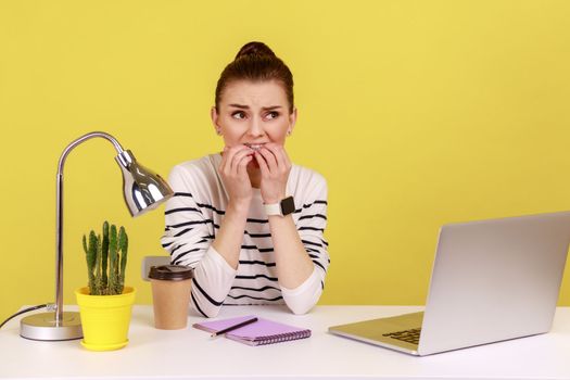 Anxious young woman office manager biting her nails sitting at workplace and looking at camera with nervous worry expression. Indoor studio studio shot isolated on yellow background.