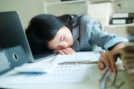 Young asian Business woman tired woman sleeping on desk with laptop at workplace alone, napping on working desk in front of her laptop in office, overworked with head on desk at the office.