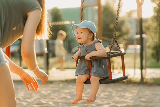 Toddler baby girl on a swing on the warm summer evening. Mother is swinging her young daughter on a sunny playground.