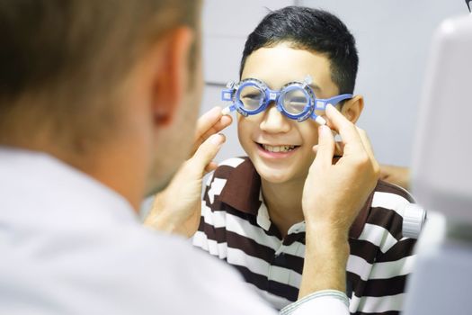 Smart young boy sitting in optometrist cabinet having his eyesight checking, examining, testing with trial frame glasses by professional optician for new pairs of eyeglasses.