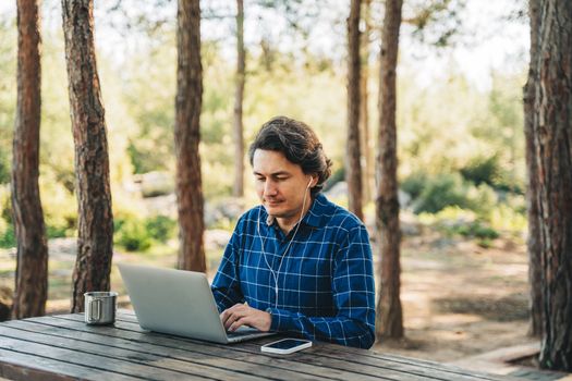 Happy cheerful man with a laptop sitting outdoors in nature forest park. Freelancer with computer typing, blogging, browsing in vacation in mountains. Freelance, distance work concept.