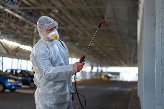 Worker wearing protective suit disinfection gear disinfect surface public place parking. Side view of disinfector safety overall and respiratory mask Covid pandemic disinfection preventive measures