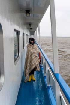 a woman wrapped in a plaid walks along the side of a ship sailing on the sea, a woman in a hood looks at the waves. Gray waves and a gray overcast sky. floating ship