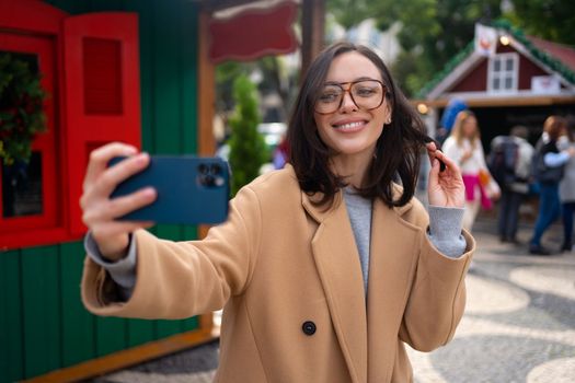 Woman taking selfie by smartphone camera outside. Caucasian female tourist in eyeglasses smiling and making selfie on European city street autumn sunny day dressed stylish coat