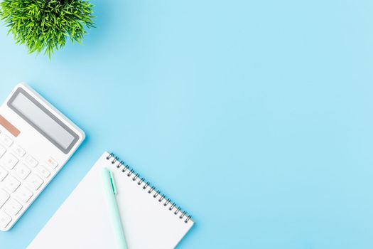 Spiral notebook with white calculator pen and green plant on a blue background. Business concept, tax calculation, office work or freelance work. Flat lay.