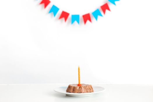 A white table with a small canned animal cake on a white saucer and an orange candle. In the background, multi-colored flags hang. Pet birthday concept. Copy space