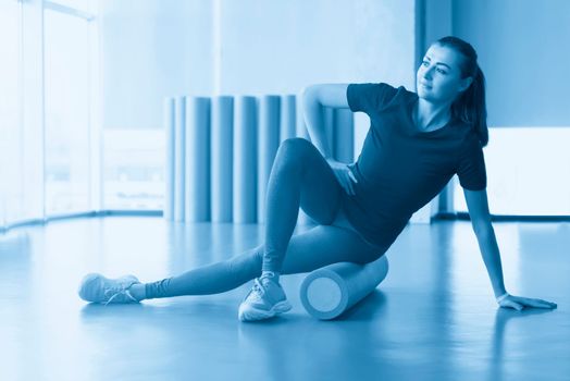 Attractive female doing foam roller exercise and posing in modern bright fitness center. Toned image.