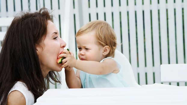 summer, in the garden, a funny one-year-old blonde girl treats her mother with watermelon, feeds her from her hands, the girl also eats watermelon. High quality photo